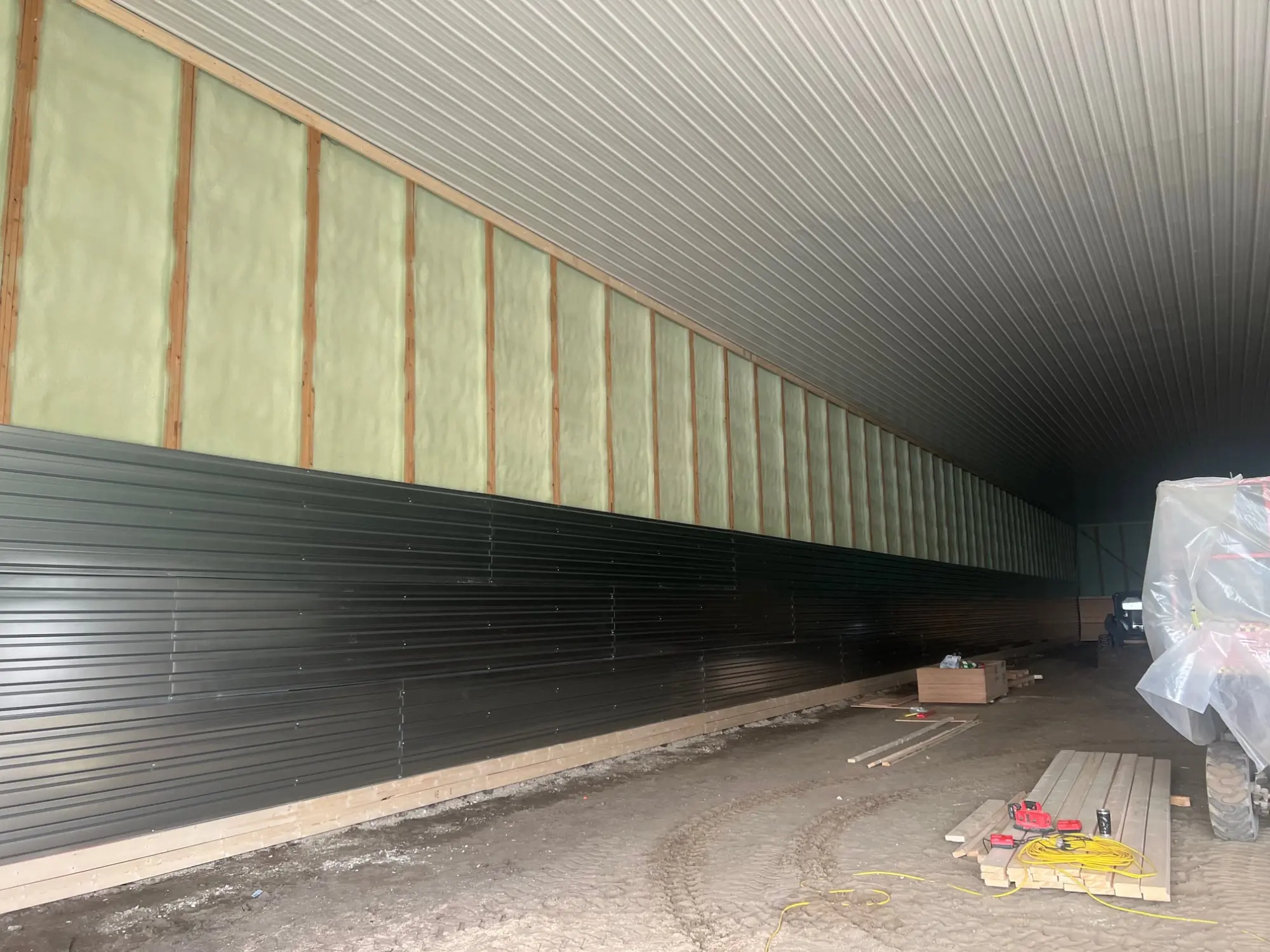Long commercial wall with closed-cell foam in the upper studs and dark metal siding at the base, dirt floor and stacked lumber.