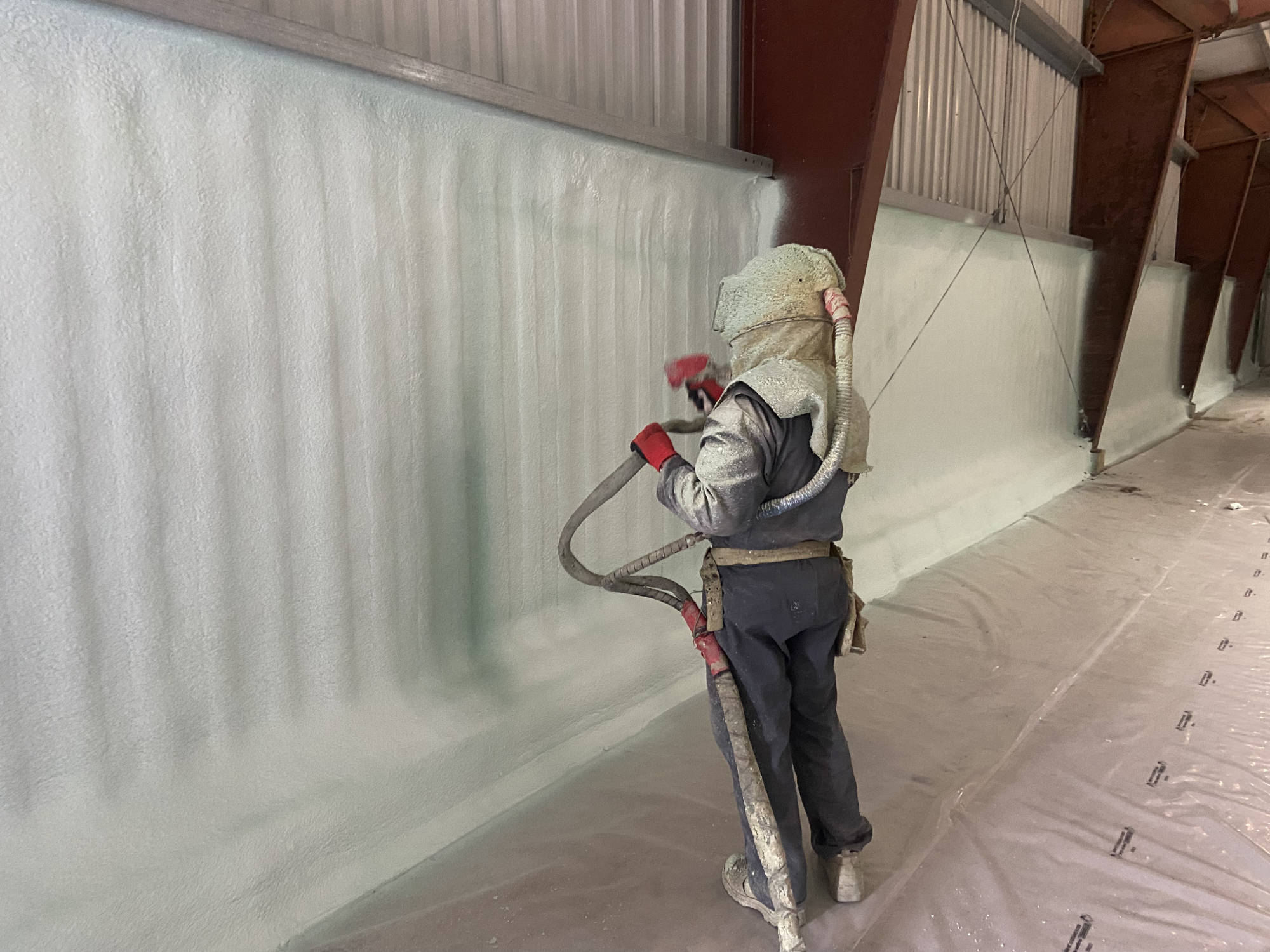 Sprayer in a full hood and grey suit applying closed-cell foam to a corrugated metal wall, plastic on the floor.