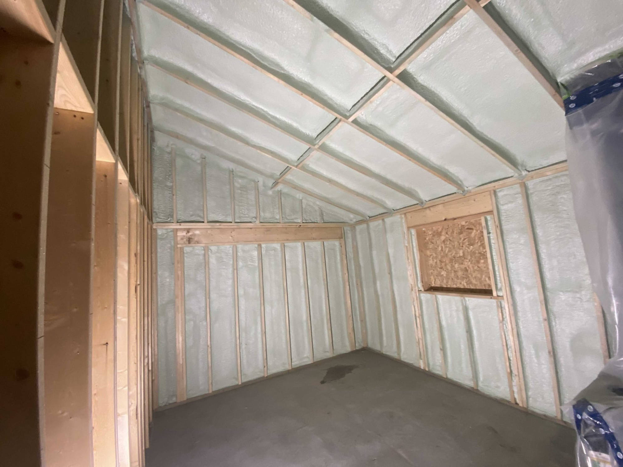 Wood-framed room with closed-cell spray foam in the walls and sloped ceiling, concrete floor and a boarded window.