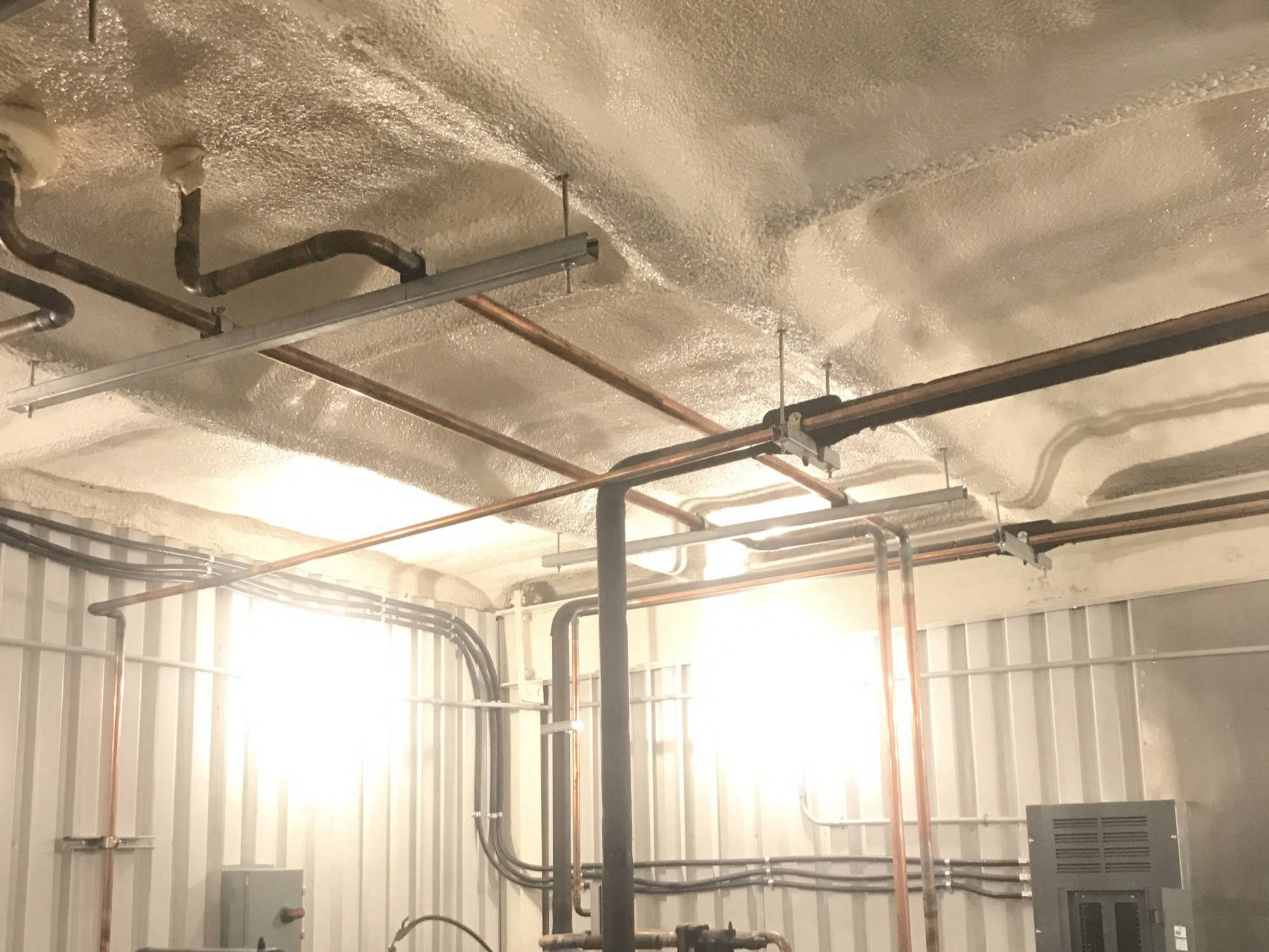 Closed-cell spray foam on a mechanical-room ceiling, sprayed around copper pipes, conduits and hangers.
