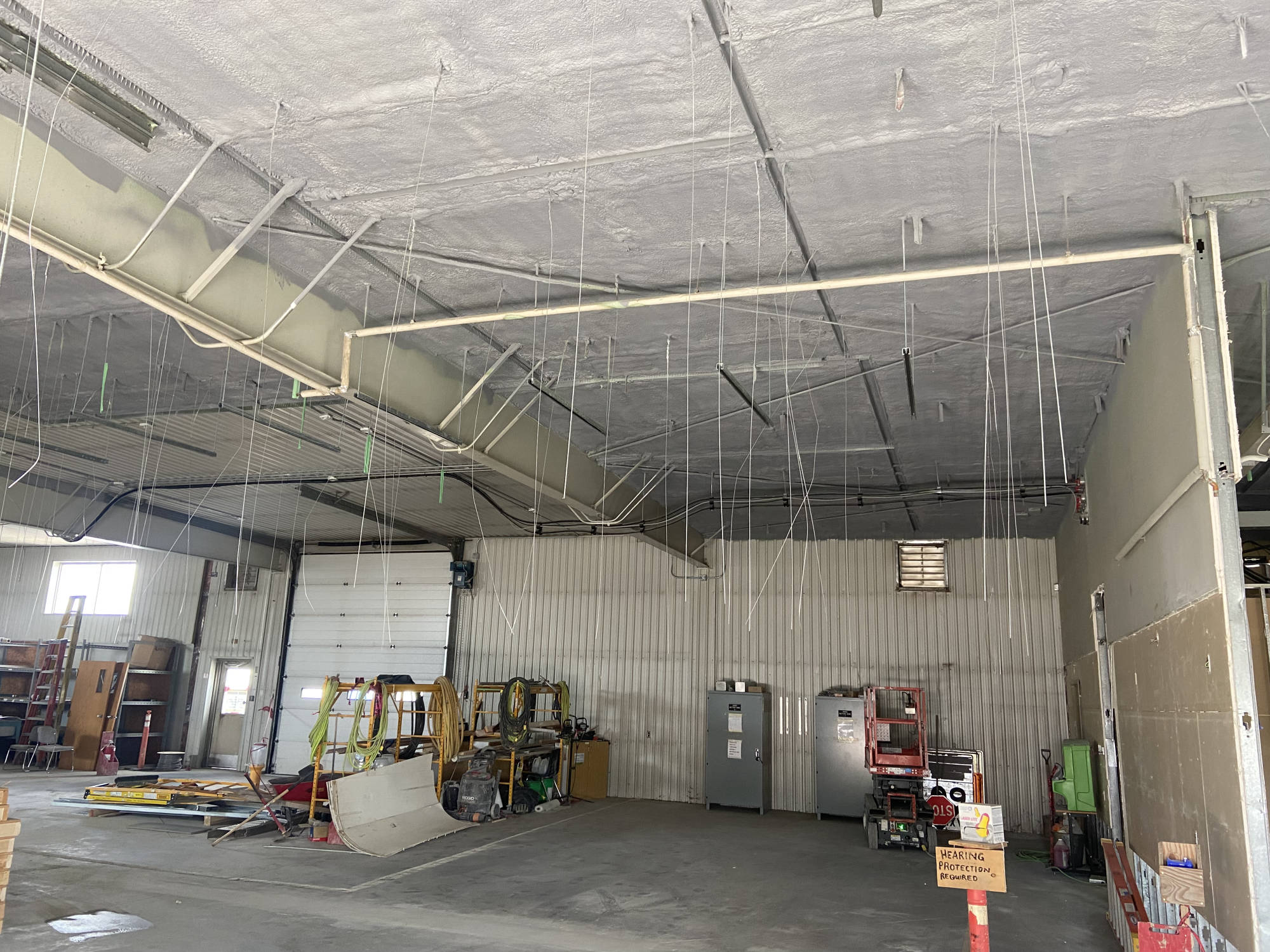 Industrial shop interior with a spray-applied coating on the ceiling, corrugated walls and a hearing-protection sign.