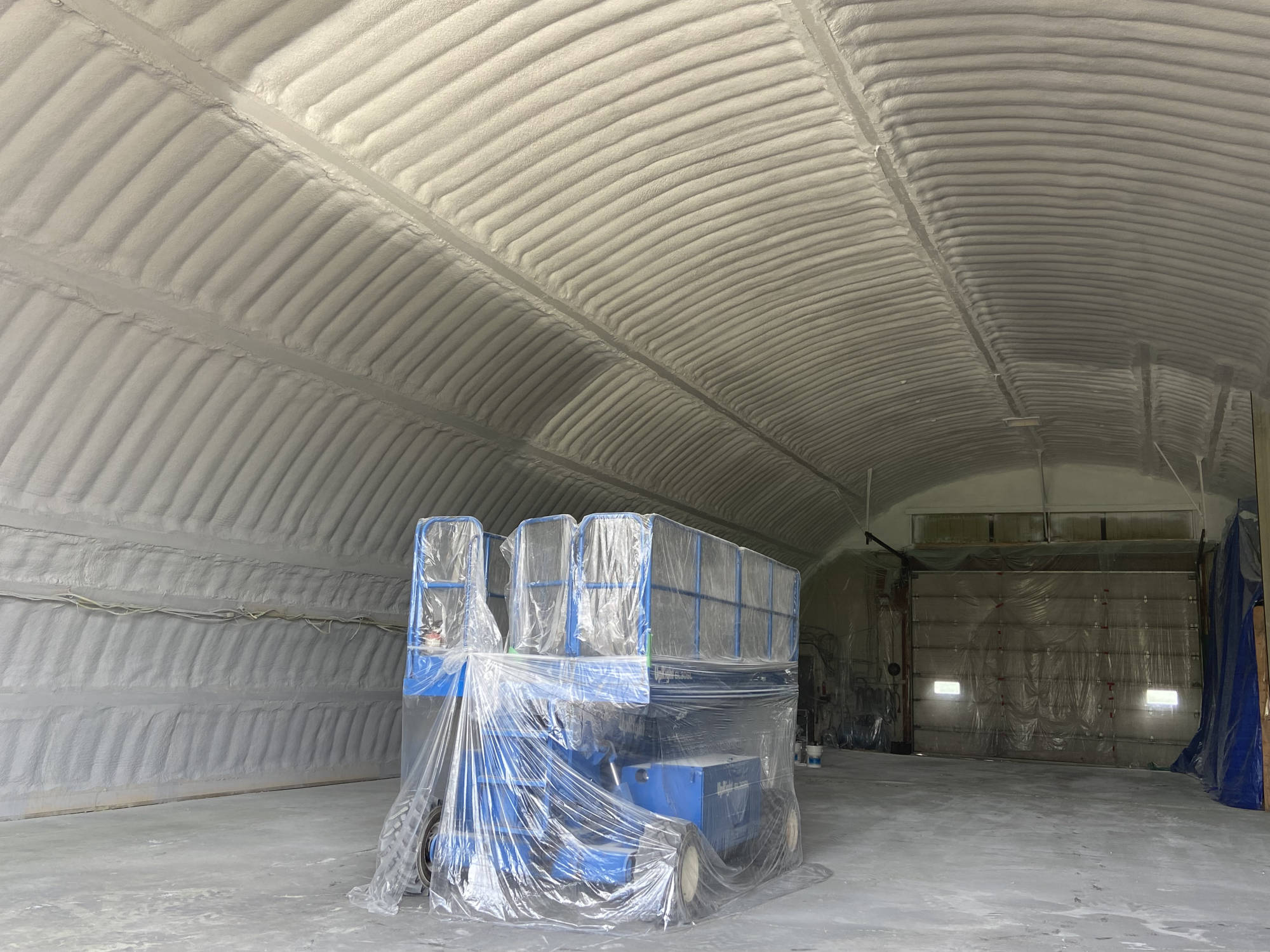 Arched building interior fully foamed, with a scissor lift wrapped in plastic on the concrete floor.