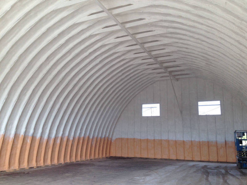 Arched metal building with white foam on the upper shell and a tan coating along the base.