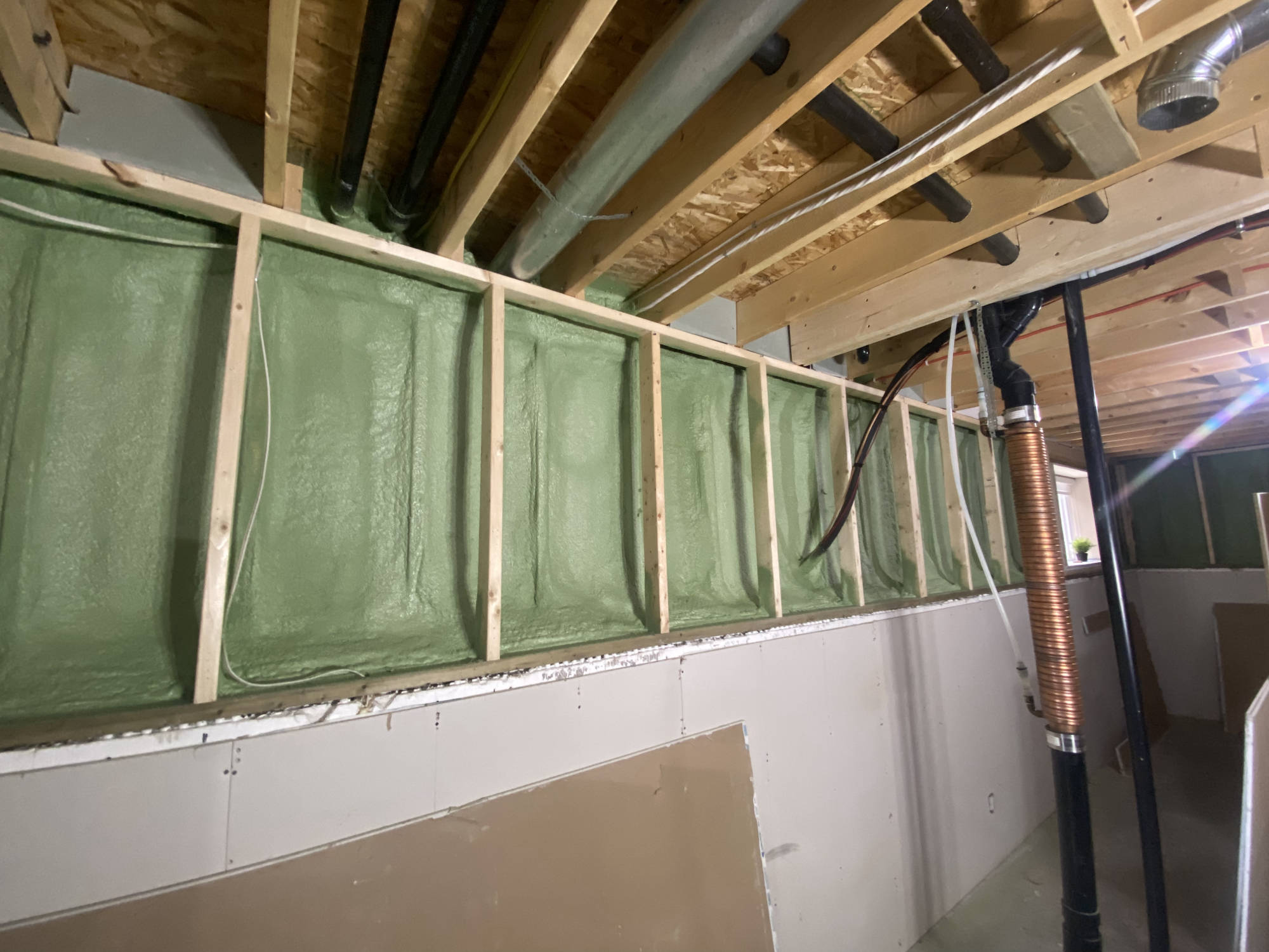 Closed-cell spray foam in basement wall studs under floor joists, with ductwork and plumbing above.