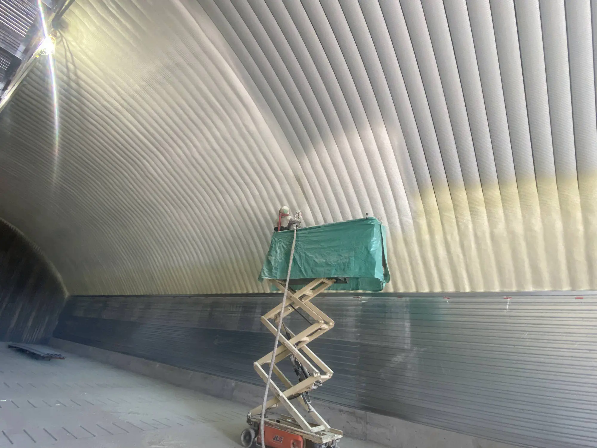 Sprayer on a scissor lift applying closed-cell foam to the arched ceiling of a large metal building.