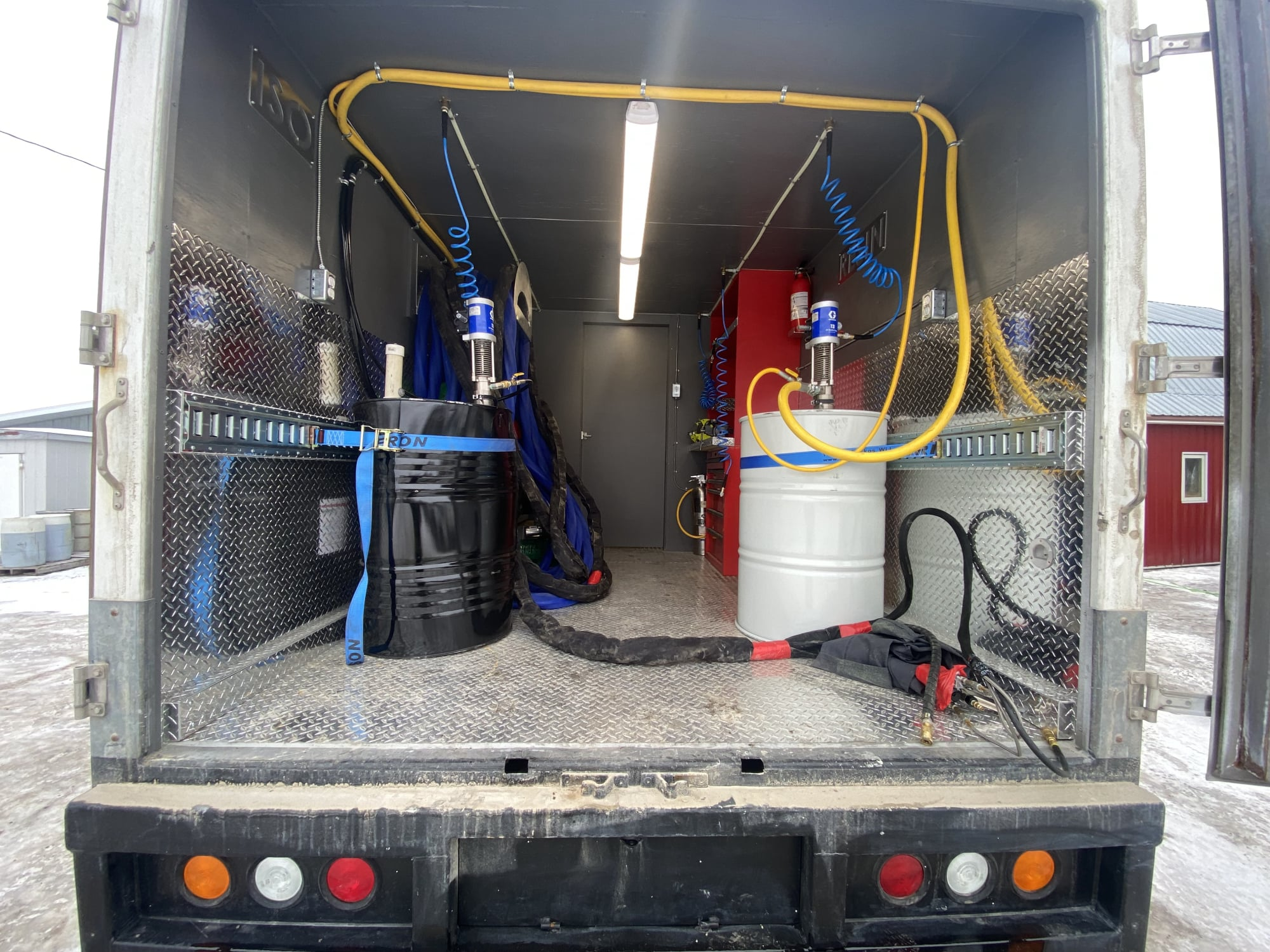 Looking into a spray foam trailer: black and white drums with transfer pumps, diamond-plate floor and coiled hose.