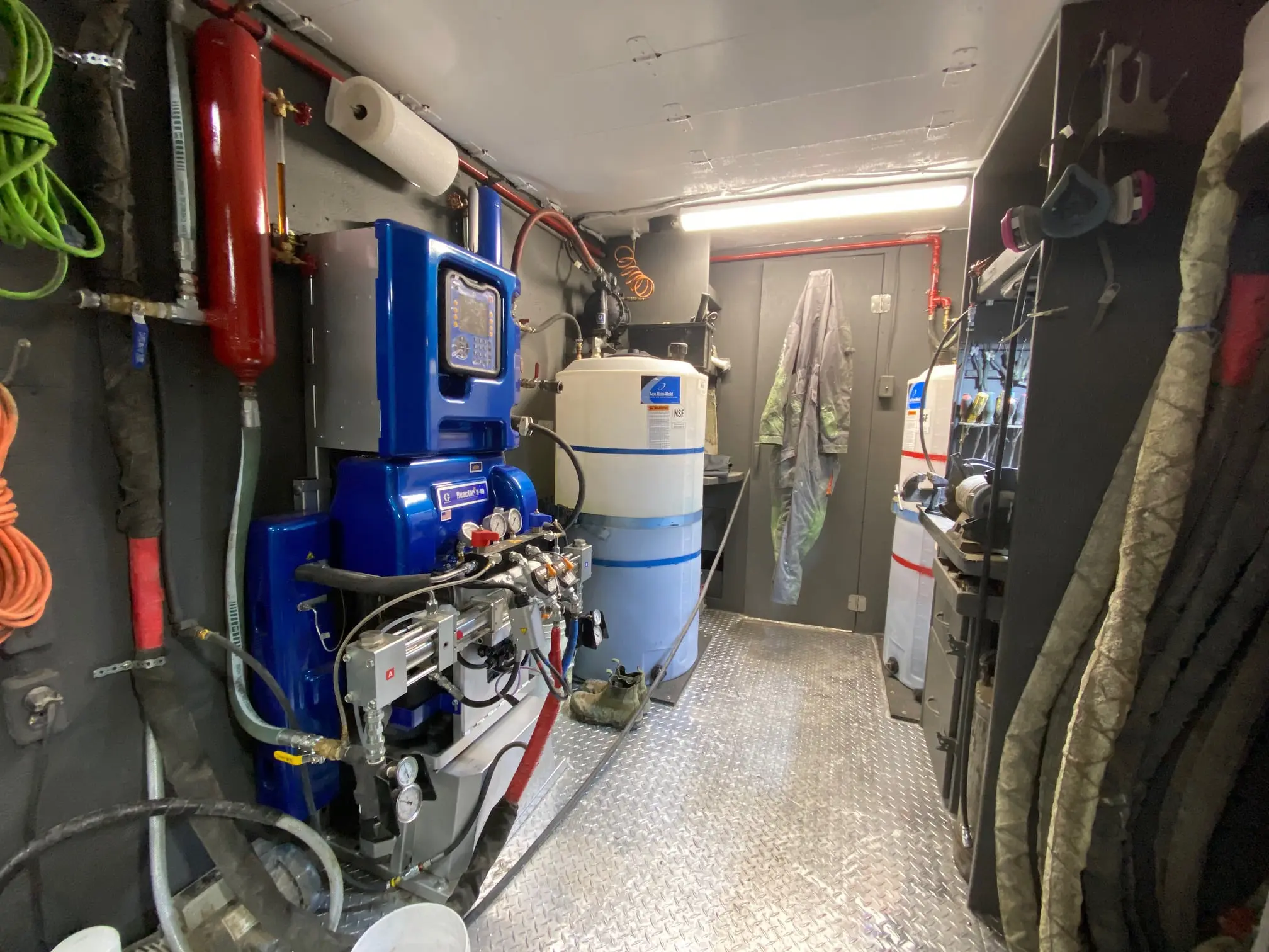 Interior of a spray foam rig with a blue Graco Reactor proportioner, drums, hoses and a coiled gun line.